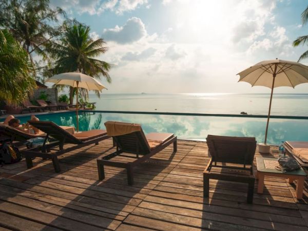 A serene deck overlooking a calm ocean with lounge chairs, parasols, and a refreshing pool, surrounded by lush palm trees under a partly cloudy sky.