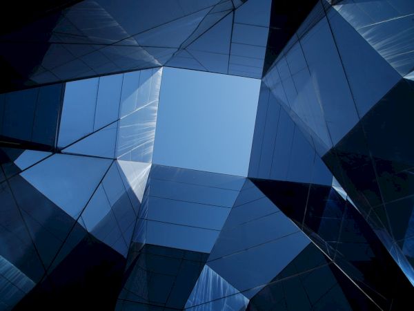 The image shows a geometric arrangement of reflective glass panes forming a modern architectural structure, with a clear sky visible in the center.