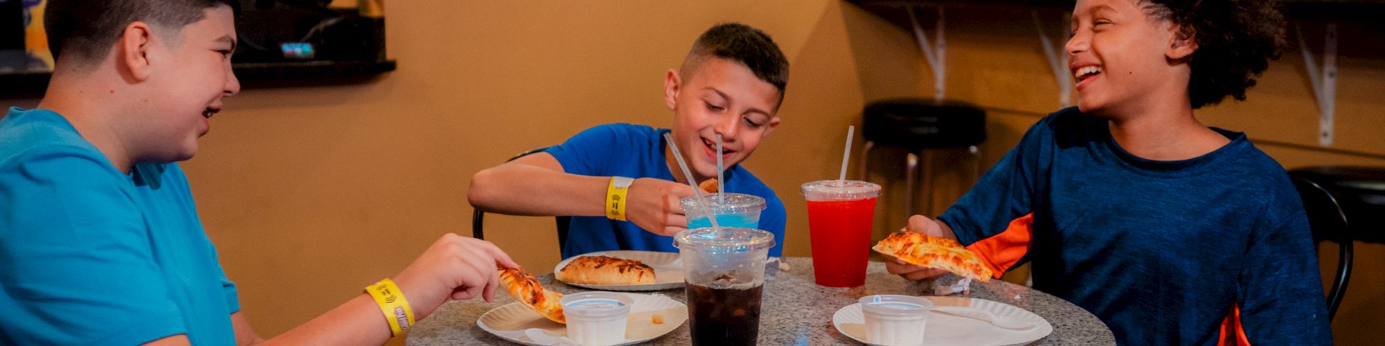 Three kids are sitting at a table, eating pizza and drinking soda, and sharing a laugh in what looks like a casual dining setting.