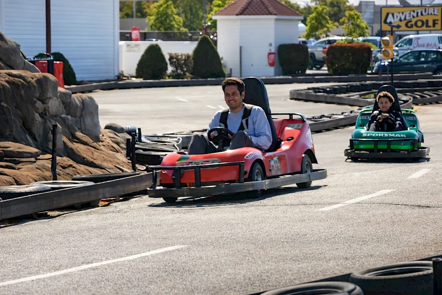 Two people drive go-karts on a paved track; a red kart leads a green one while spectators and a rocky barrier line the course.