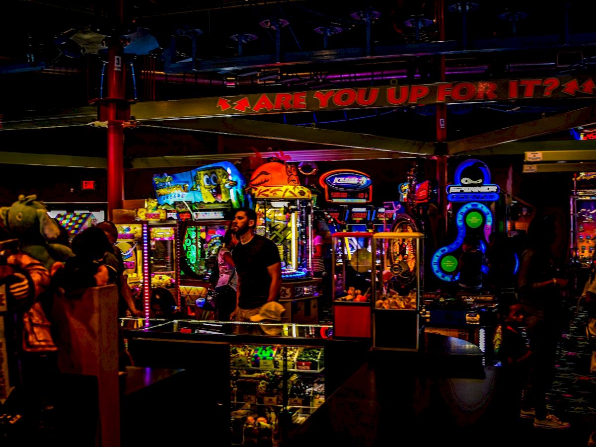 The image shows a dimly lit arcade with colorful games and machines, and a person standing near them. A sign says 