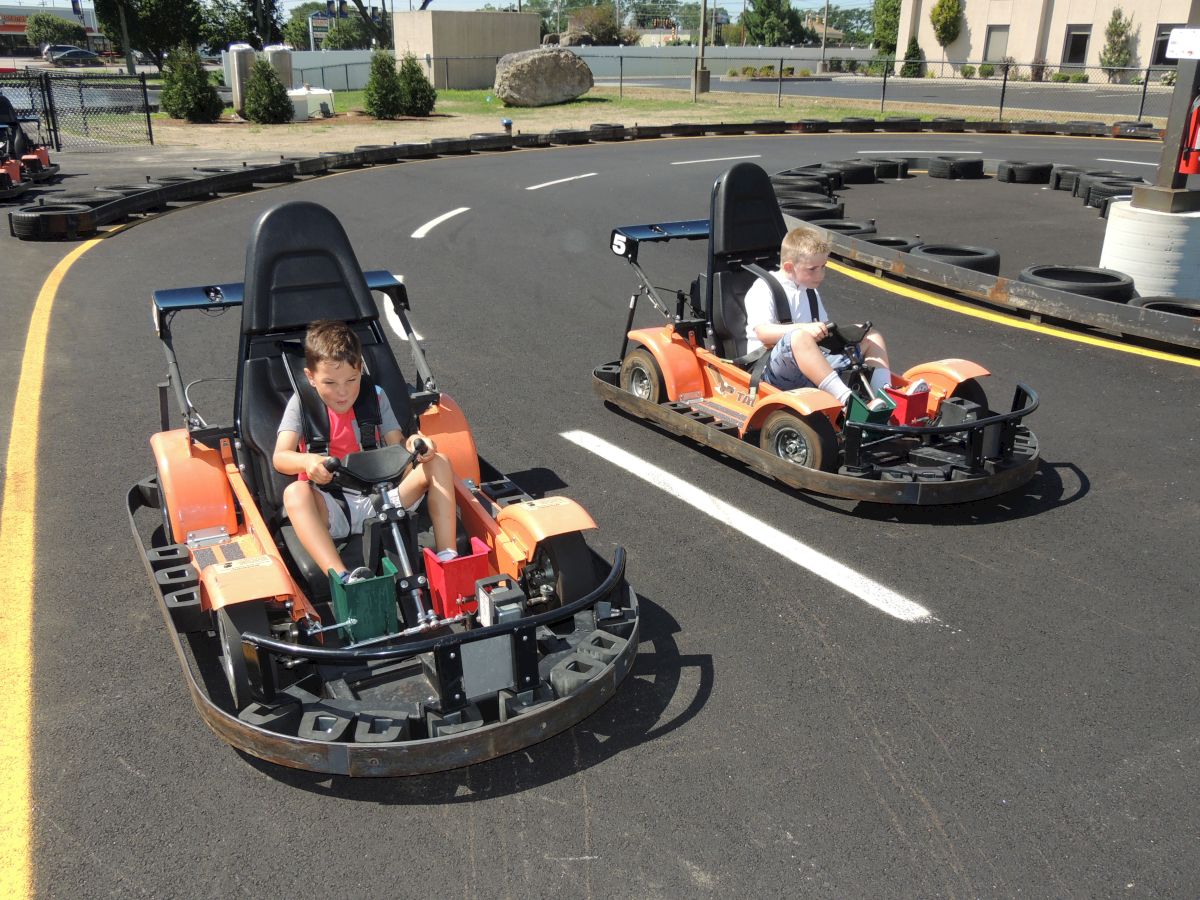 Two children driving go-karts on a track with black tires and a curve in the background, both wearing seatbelts and racing helmets.