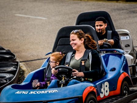 A woman and a child are driving a blue kart with the number 38, while a man follows behind in another kart on a go-kart track.