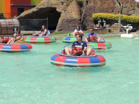 People are enjoying a sunny day in bumper boats at an amusement park, with a rocky backdrop and colorful surroundings.