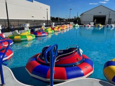 The image shows a pool filled with colorful bumper boats, situated outdoors with a building and clear blue sky in the background.