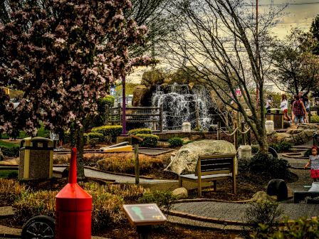 A park scene with a fountain, benches, lush trees, and blooming flowers, along with people enjoying the surroundings.