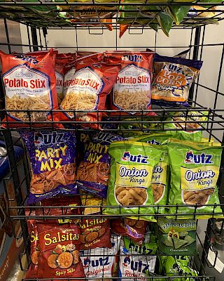 The image shows a variety of snack bags on a metal rack, including Potato Stix, Party Mix, Onions, and Salsitas chips.