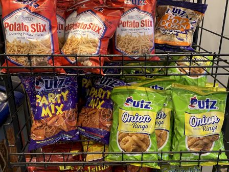 The image shows a variety of snack bags on a metal rack, including Potato Stix, Party Mix, Onions, and Salsitas chips.