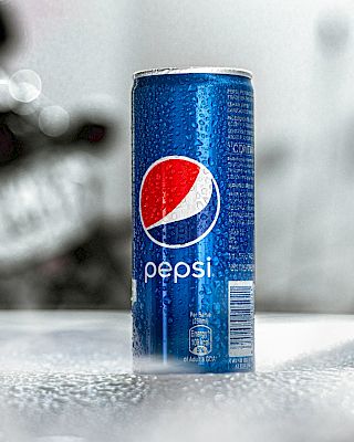 The image shows a blue can of Pepsi placed on a wet surface with a blurred and out-of-focus background.