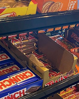 The image shows a display of various candy bars on store shelves, including Crunch, Twix, Rolo, and Reese's.