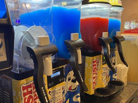 The image shows a row of slushie machines with blue and red slush flavors, labeled 