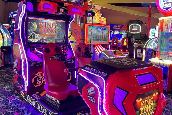 This image shows an arcade game machine with two seats and multiple screens in a dimly lit arcade with colorful lights and other games in the background.