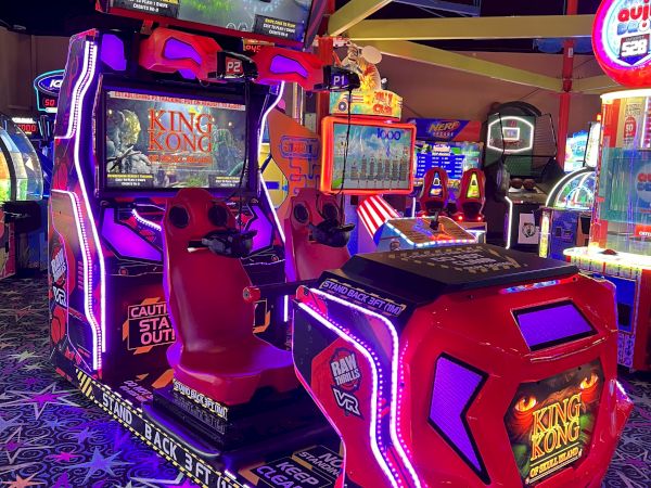 This image shows an arcade game machine with two seats and multiple screens in a dimly lit arcade with colorful lights and other games in the background.