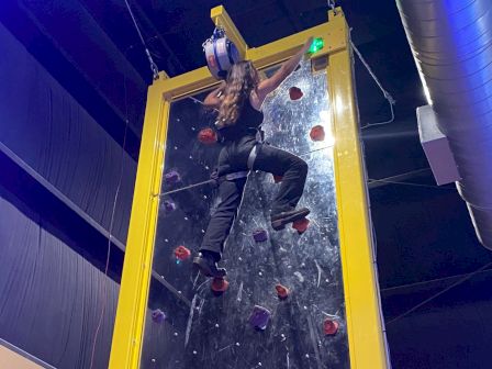 A person is climbing an indoor climbing wall, reaching near the top and preparing to touch a green button, with safety gear on.