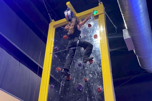 A person is climbing an indoor climbing wall, reaching near the top and preparing to touch a green button, with safety gear on.