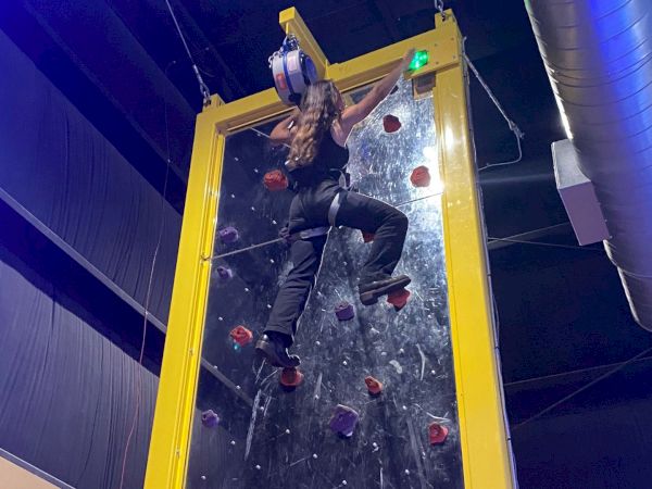 A person is climbing an indoor climbing wall, reaching near the top and preparing to touch a green button, with safety gear on.