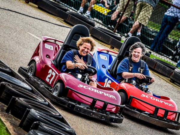 Two people are racing in colorful go-karts on a track, smiling and having fun.