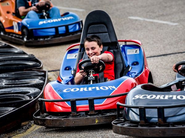 A person is driving a red go-kart labeled "SCORPION" on a track with tire barriers, with other go-karts in the background, ending the sentence.