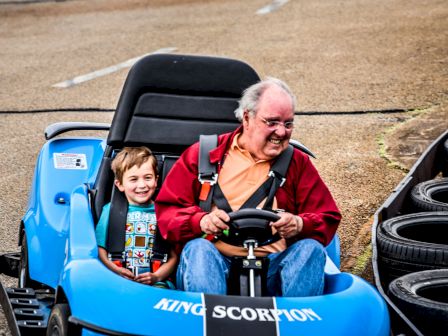 A smiling child and older man in a red jacket drive a blue "King Scorpion" go-kart on a track beside stacked tires.