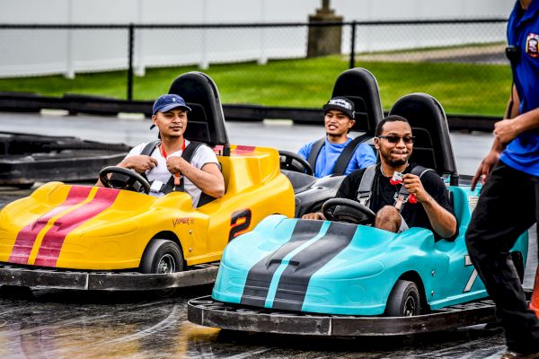 People driving yellow and blue go-karts on a track, enjoying a fun time. Another person is partially seen on the right.