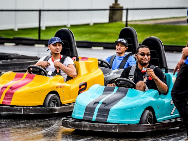 People driving yellow and blue go-karts on a track, enjoying a fun time. Another person is partially seen on the right.