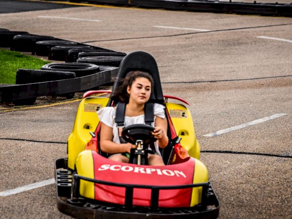 A person is driving a yellow and red go-kart on a racetrack, marked with "Scorpion" on the front, wearing a white top and showing a focused expression.