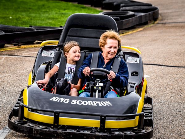 Two people enjoy a ride in a yellow go-kart labeled "KING SCORPION" on a race track, both smiling and looking happy.