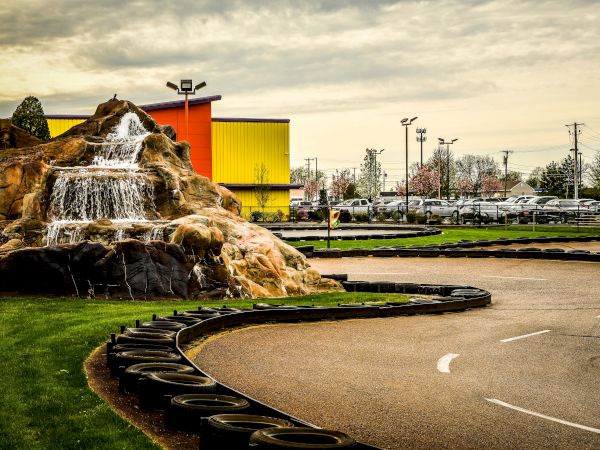 A go-kart track with a curve bordered by tires, adjacent to a rock formation with a waterfall, near buildings and trees under a cloudy sky.