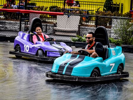 Two people are enjoying a ride in mini electric cars at a fun amusement park attraction, with big smiles on their faces as they drive around.