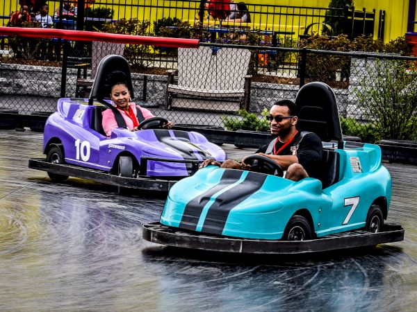 Two people are enjoying a ride in mini electric cars at a fun amusement park attraction, with big smiles on their faces as they drive around.