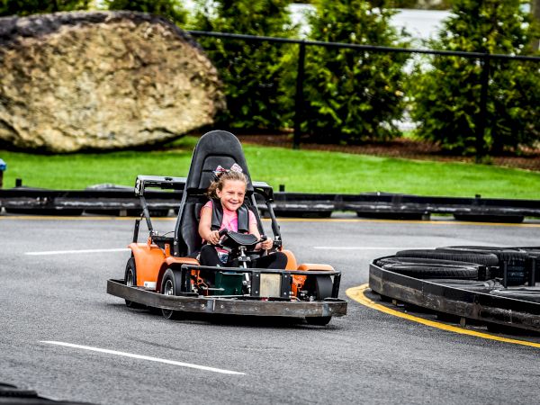 A child is driving a go-kart on an outdoor track, smiling and having fun, while wearing a safety harness and surrounded by greenery and tires.