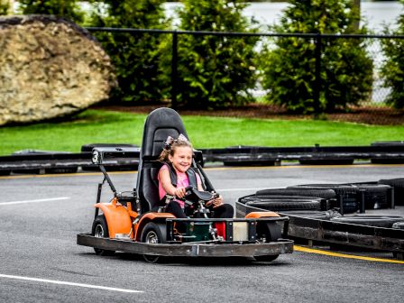A child is driving a go-kart joyfully on an outdoor track with greenery and a rock in the background.