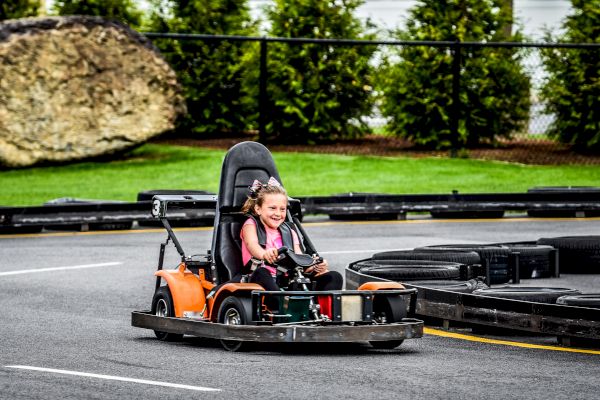 A child is driving a go-kart joyfully on an outdoor track with greenery and a rock in the background.