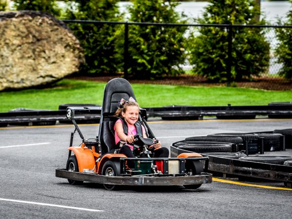 A child is driving a go-kart joyfully on an outdoor track with greenery and a rock in the background.