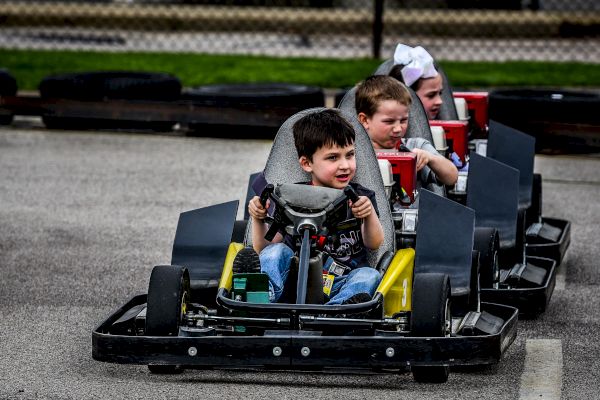 Three children are driving go-karts outdoors, with the leading child smiling and focused.