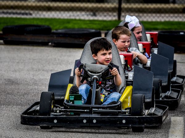 Three children are driving go-karts outdoors, with the leading child smiling and focused.