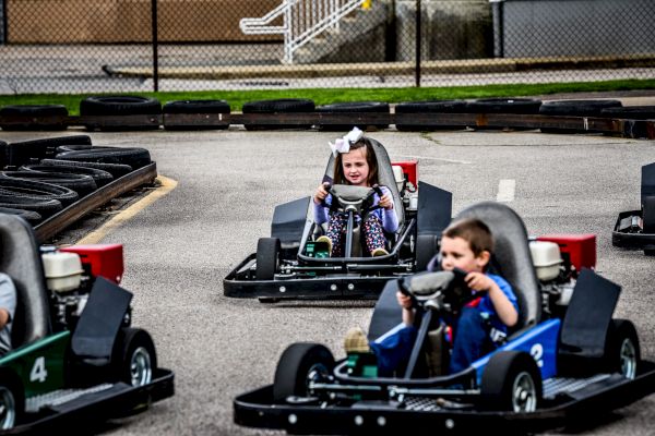 Children are driving go-karts on a track, having fun and smiling, with safety barriers on both sides to ensure a secure environment.