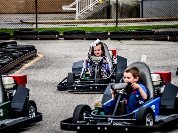 Children are driving go-karts on a track, having fun and smiling, with safety barriers on both sides to ensure a secure environment.