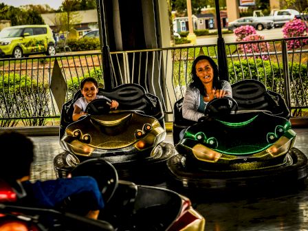 People enjoying a ride in bumper cars on an amusement park track, with a fence and greenery in the background.