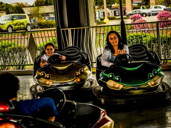 People enjoying a ride in bumper cars on an amusement park track, with a fence and greenery in the background.
