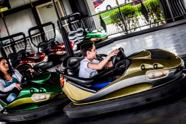 Two people are enjoying a ride on bumper cars at an amusement park, one in a green car and the other in a gold car, with colorful lights in the background.