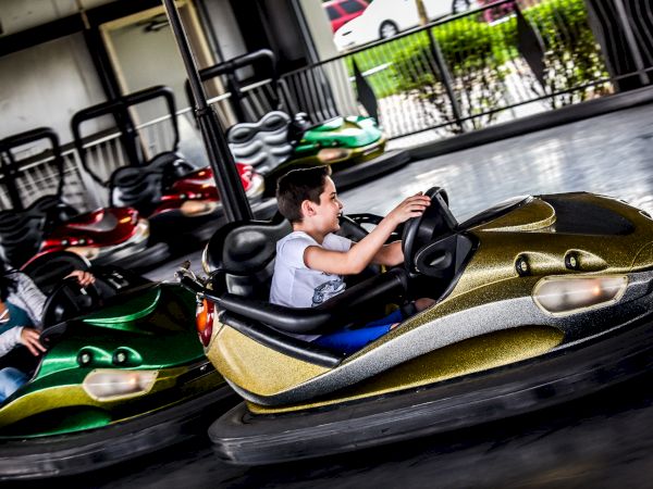 Two people are enjoying a ride on bumper cars at an amusement park, one in a green car and the other in a gold car, with colorful lights in the background.