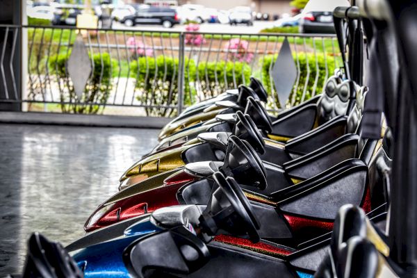 Image shows a row of parked bumper cars, with various colors visible, in an amusement park setting, with greenery and cars in the background.