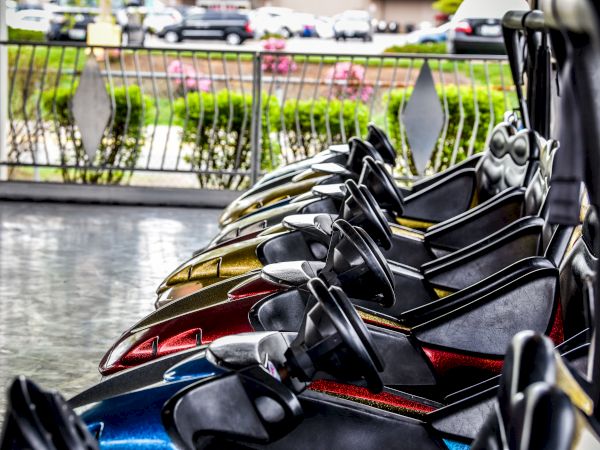 Image shows a row of parked bumper cars, with various colors visible, in an amusement park setting, with greenery and cars in the background.