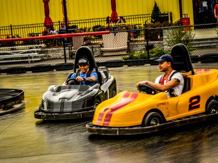 Two people are driving go-karts on a track, one in a gray kart and the other in a yellow kart numbered 2.