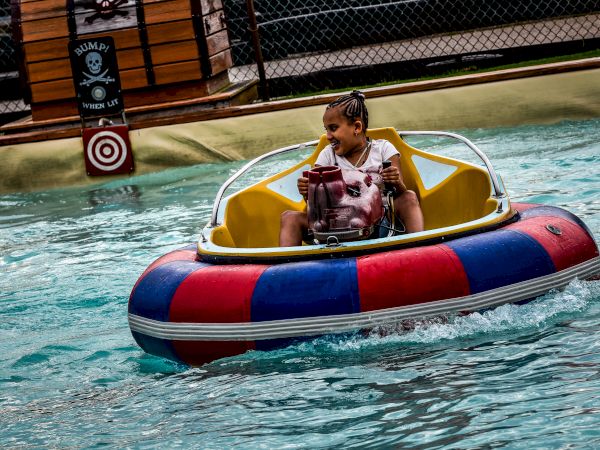 A person is riding a bumper boat in a pool, near a sign with a target and a "Yummy Pirate" logo in the background.