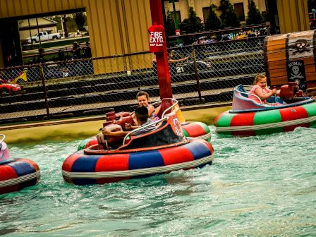 People are riding colorful bumper boats in a pool at an amusement park or similar setting, with water splashing around them.