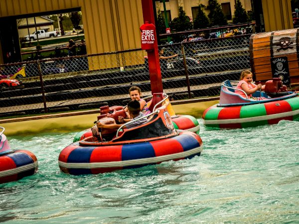 People are riding colorful bumper boats in a pool at an amusement park or similar setting, with water splashing around them.