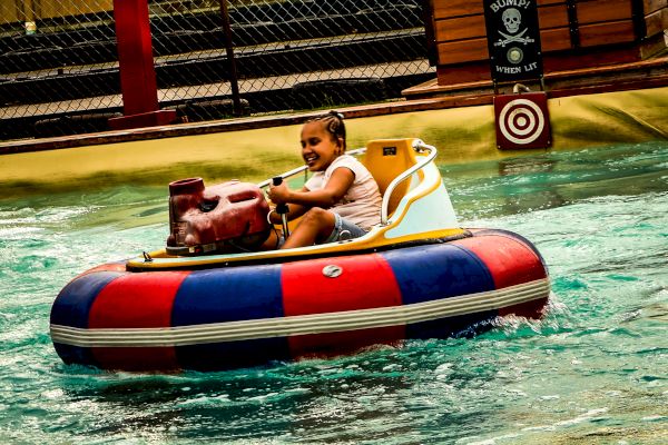 A child is enjoying a ride on a circular bumper boat in a water amusement park or pool. The child is smiling and holding the steering controls.