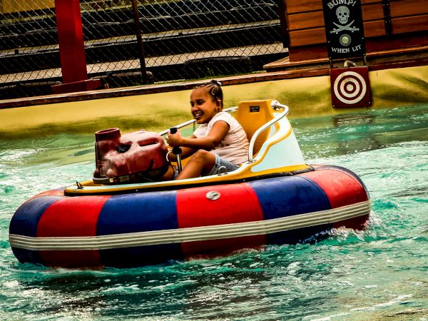 A child is enjoying a ride on a circular bumper boat in a water amusement park or pool. The child is smiling and holding the steering controls.